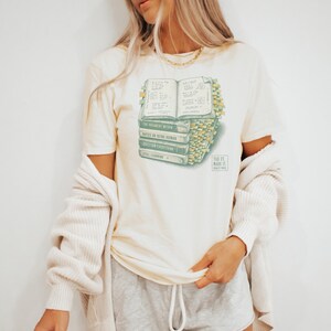 May include: A cream-colored short-sleeve t-shirt featuring a graphic of a stack of books with numerous yellow sticky tabs. The books have titles including The Argument Within, Notes on Being Human, Question Everything, and Still Learning.