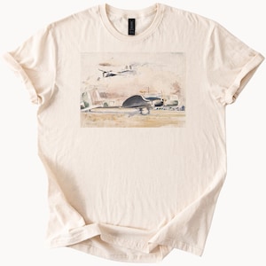 May include: Cream-colored t-shirt featuring a watercolor painting of two vintage airplanes. The artwork depicts a larger plane on the ground and a smaller one in the sky, set against a neutral background. The shirt is made of soft fabric.