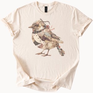 May include: Cream-colored t-shirt featuring a detailed illustration of a bird. The bird is composed of various fabric patches in shades of brown, beige, and cream, with accents of black and red. The bird has a decorative ribbon and a small rose.