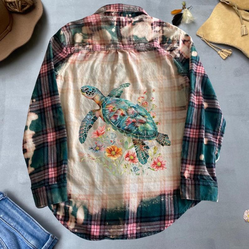 May include: A green and pink plaid flannel shirt with a bleached effect. The shirt features a graphic of a sea turtle with flowers on the back.