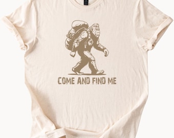 May include: A beige t-shirt with a brown illustration of a Bigfoot carrying a backpack and the text "Come and find me".