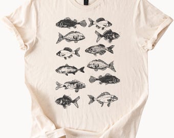 Könnte beinhalten: Cremefarbenes T-Shirt mit einem sich wiederholenden Muster aus verschiedenen Fischillustrationen in schwarzer Tinte. Die Fische sind in verschiedenen Größen und Ausrichtungen dargestellt, was ein visuell interessantes Design ergibt. Das Shirt hat kurze Ärmel und einen Rundhalsausschnitt.