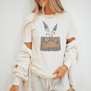 May include: A cream-colored short-sleeve t-shirt featuring a graphic print of a weathered, rectangular sign with a screaming face and the text CLOSED PERMANENTLY, topped with two purple flowering stems. The shirt is worn with a beige cardigan.