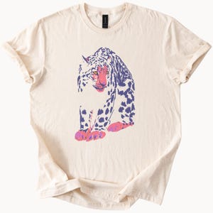 以下が含まれることがあります： クリーム色のTシャツ。紫、ピンク、白の色合いで描かれた、様式化されたヒョウのデザインが特徴です。ピンク色の顔と足を持っています。Tシャツは半袖で、クルーネックです。