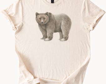May include: Cream-colored t-shirt featuring a detailed illustration of a grizzly bear. The bear is depicted in shades of gray and brown, standing upright. The shirt has a classic crew neck and short sleeves.