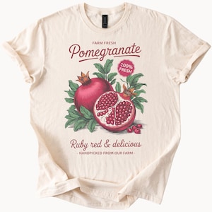 Może przedstawiać: Beżowa koszulka z nadrukiem granatu. Na koszulce widnieje tekst "FARM FRESH Pomegranate 100% FRESH Ruby red & delicious - HANDPICKED FROM OUR FARM - ".