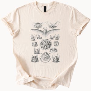 May include: A cream-coloured t-shirt displaying a detailed black and white illustration of various bat species. The design includes multiple bat faces and full body depictions, with scientific labels. A classic crew neck tee.