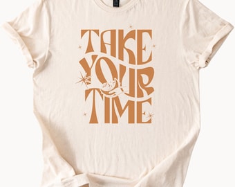 May include: Cream-coloured t-shirt featuring the words "TAKE YOUR TIME" in an orange retro font. The design includes a star and a cat holding a crescent moon. A casual, comfortable tee.