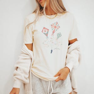 Può includere: T-shirt color crema con una grafica di tre aquiloni rosa, rossi e turchese, con disegni floreali e geometrici. Gli aquiloni sono collegati da fili e tenuti da una mano. Il testo "north wind" è sotto.