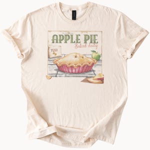 Puede incluir: Camiseta color crema con un gráfico de tarta de manzana y el texto "APPLE PIE Baked daily". El gráfico incluye una tarta, una manzana y una bolsa de harina. La camiseta tiene mangas cortas y cuello redondo.