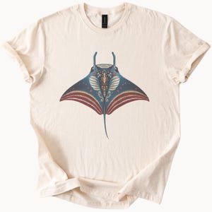 May include: Cream-colored t-shirt featuring a detailed illustration of a manta ray. The ray is depicted in shades of blue, red, and gold, with intricate patterns. The shirt has short sleeves and a crew neck.