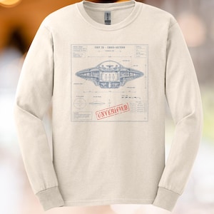 May include: A beige long-sleeve crewneck shirt featuring a blue technical blueprint graphic of a UFO cross-section. The print includes the text UNIT 7D - CROSS-SECTION and a red stamp that reads UNVERIFIED, along with various technical labels.