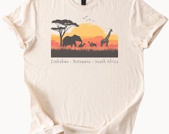 May include: Cream-colored t-shirt featuring a sunset scene with African wildlife silhouettes. An elephant, giraffes, and other animals are depicted against a backdrop of a setting sun. The text "Zimbabwe - Botswana - South Africa" is printed below the image.