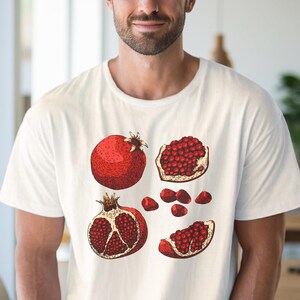 May include: White t-shirt featuring a detailed illustration of pomegranates. The design includes a whole pomegranate, sections, and individual seeds in shades of red and brown. The shirt is a classic crew neck.