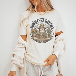 May include: Cream-colored t-shirt featuring a circular graphic with the text "Support Your Local Bees." The graphic includes a cartoon bee wearing an apron, surrounded by flowers and mugs. The phrase "Made with Love" is also present.