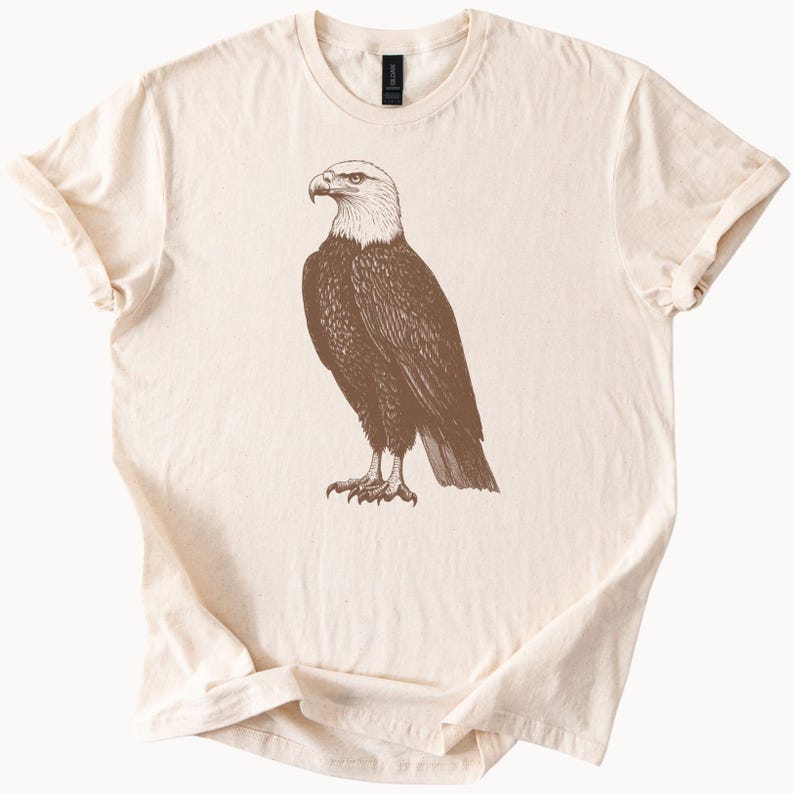 May include: A beige t-shirt with a brown illustration of a bald eagle on the front.