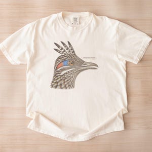 May include: An off-white short-sleeve t-shirt featuring a detailed illustration of a roadrunner head in profile. The bird has brown and tan feathers, a blue and red patch near the eye, and the text ORBITAL PATCH printed in small black letters.