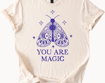 May include: A cream-colored t-shirt with a blue graphic of a moth with moon and star details. The text "YOU ARE MAGIC" is printed below the moth.