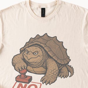 Pode incluir: Uma t-shirt de cor creme com uma ilustração gráfica de uma tartaruga-aligátor castanha com uma expressão severa. A tartaruga pressiona um carimbo vermelho sobre uma superfície, com o texto parcial vermelho NO visível abaixo do carimbo.