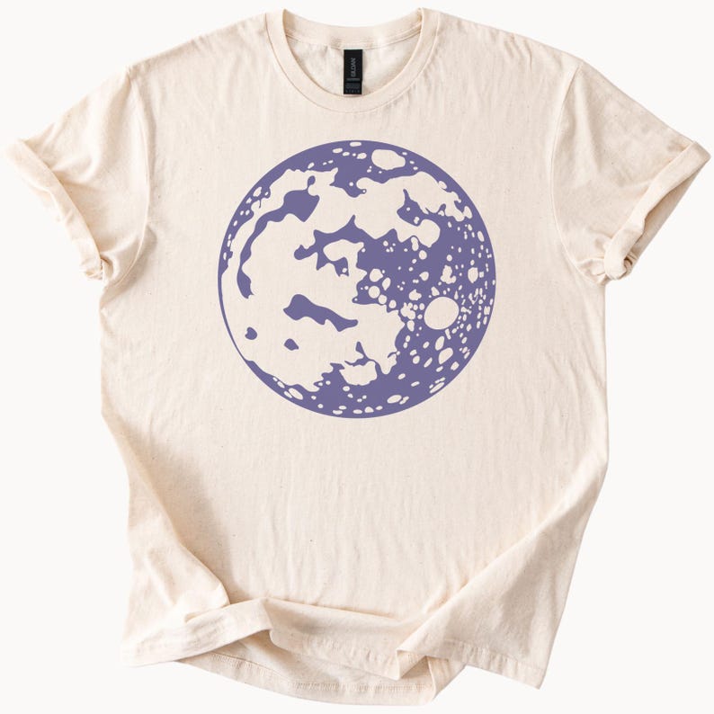 Op de afbeelding: Een licht beige t-shirt met een paarse maanprint. De maan is een gestileerde illustratie met een gedetailleerd oppervlak.