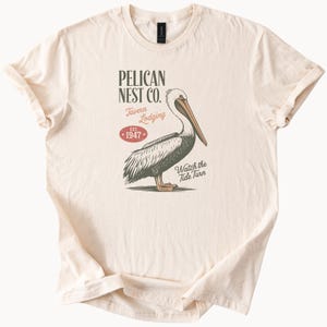 May include: A cream-coloured t-shirt with a pelican illustration and text. The design includes "PELICAN NEST CO. Tavern Lodging EST. 1947 Watch the Tide Turn". The graphic is in a vintage style. The t-shirt has rolled-up sleeves and is made of a soft material.