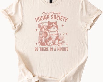 May include: A beige t-shirt with a pink graphic of a cat wearing a backpack and sunglasses. The text on the shirt reads "Out of Breath Hiking Society" and "Be There in a Minute".