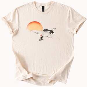 May include: A beige t-shirt with a black and white illustration of a crane flying in front of a red and orange sun.