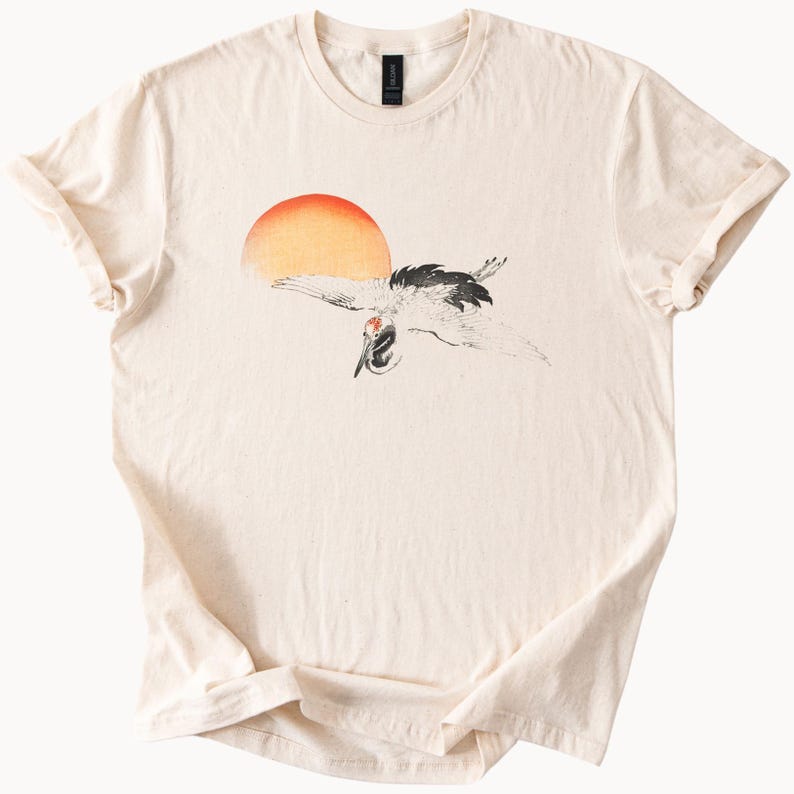 May include: A beige t-shirt with a black and white illustration of a crane flying in front of a red and orange sun.