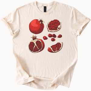 May include: Cream-colored t-shirt featuring a detailed illustration of pomegranates. The design includes whole and sliced pomegranates, showcasing the red fruit and seeds. The artwork has a vintage, botanical style.