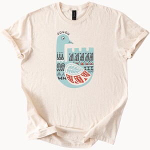 May include: A cream-coloured t-shirt with a stylised bird design in blue, red, and white. The bird has a long neck, a decorative crest, and a patterned body. The shirt is made of a soft material and has rolled-up sleeves.