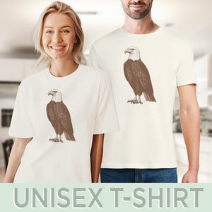 May include: Two white t-shirts with a brown bald eagle graphic printed on the front. The eagle is facing right and has a white head and tail feathers.
