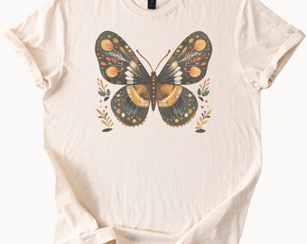 Peut inclure: T-shirt crème avec un motif de papillon détaillé. Le papillon a des ailes vert foncé avec des accents orange et jaune, et un corps marron. De petits éléments floraux entourent le papillon. Le t-shirt est en matière douce.