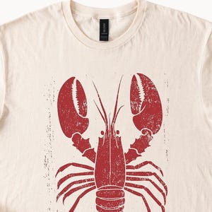以下が含まれることがあります： ベージュのTシャツに赤いロブスターのイラストが描かれています。ロブスターはヴィンテージ調で、使い古したような風合いになっています。