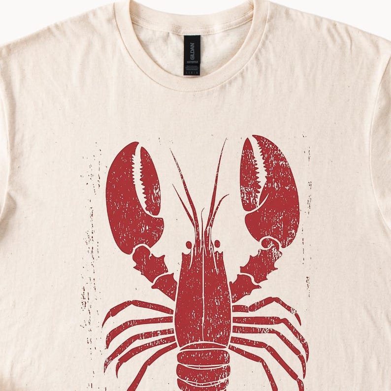 以下が含まれることがあります： ベージュのTシャツに赤いロブスターのイラストが描かれています。ロブスターはヴィンテージ調で、使い古したような風合いになっています。
