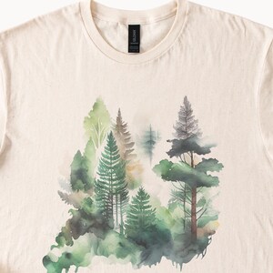 May include: A beige t-shirt with a watercolor design of a forest scene. The design features green, brown, and gray trees with a white background.