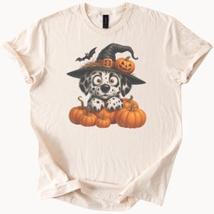 May include: Cream-colored t-shirt featuring a Dalmatian puppy wearing a witch hat with a pumpkin and bat. The puppy is surrounded by orange pumpkins, creating a Halloween theme.