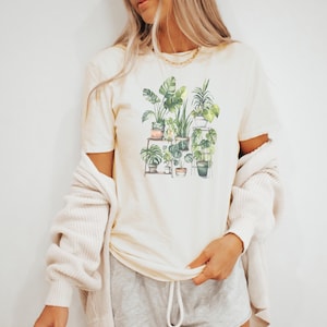 Puede incluir: Camiseta color crema con una ilustraci&oacute;n en acuarela de varias plantas en macetas. El dise&ntilde;o incluye una variedad de follaje y macetas verdes, creando un tema bot&aacute;nico. La camiseta se combina con pantalones cortos grises y una rebeca color crema.