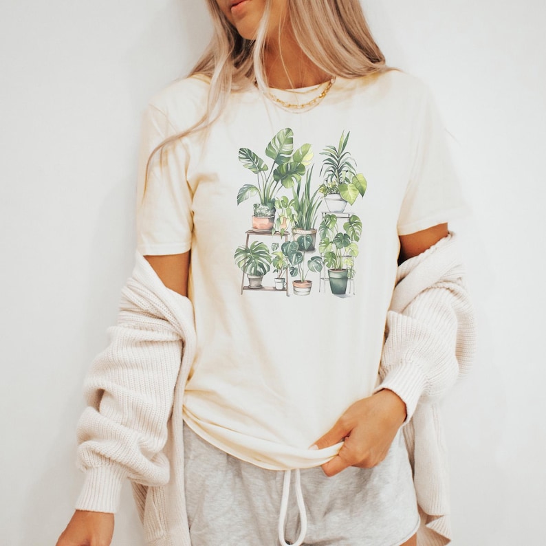 Puede incluir: Camiseta color crema con una ilustraci&oacute;n en acuarela de varias plantas en macetas. El dise&ntilde;o incluye una variedad de follaje y macetas verdes, creando un tema bot&aacute;nico. La camiseta se combina con pantalones cortos grises y una rebeca color crema.
