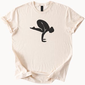 May include: Cream-colored t-shirt featuring a black silhouette of a person in a yoga pose. The design shows a person balancing on their hands, with legs extended. The shirt has a crew neck and short sleeves.