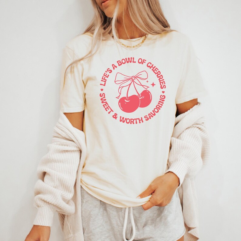 May include: A white t-shirt with a pink graphic of two cherries with a bow and the text "Life's a bowl of cherries, sweet & worth savoring."