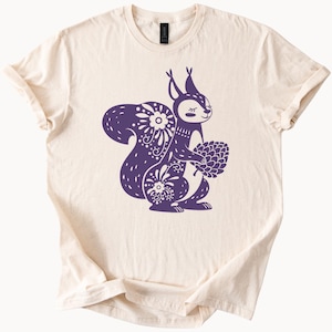 Op de afbeelding: Een crèmekleurig T-shirt met een paars eekhoornontwerp. De eekhoorn houdt een dennenappel vast en heeft een bloemenpatroon.