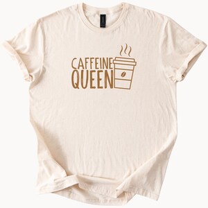 Op de afbeelding: Crèmekleurig T-shirt met de woorden "CAFFEINE QUEEN" in bruine letters, vergezeld van een bruine koffiekop-afbeelding met stoom en een koffieboon. Het shirt heeft korte mouwen en een ronde hals.