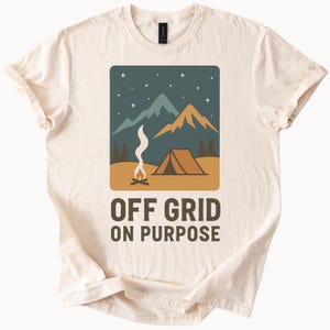 May include: A beige t-shirt with a graphic of a campfire, tent, and mountains under a starry night sky. The text "OFF GRID ON PURPOSE" is printed below the graphic.