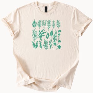 Peut inclure: T-shirt crème avec une collection d'illustrations d'algues vertes. Le motif comprend diverses formes et tailles de plantes aquatiques, créant un thème botanique. Le t-shirt a des manches courtes et un col rond.