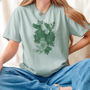 May include: A light sage green short-sleeve t-shirt featuring a dark green graphic print of a phoenix bird surrounded by detailed floral elements. The shirt has a crew neckline and a relaxed fit, showcasing a monochromatic design on the front.