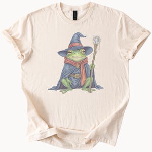 May include: Cream-coloured t-shirt with a fantasy illustration of a frog wizard. The frog is depicted in a blue wizard hat and cape, a red scarf, and holds a staff with a crystal ball. The design is centrally placed on the shirt.