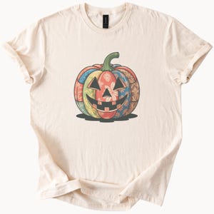 May include: Cream-colored t-shirt featuring a colorful patchwork pumpkin design. The pumpkin has a smiling jack-o'-lantern face and is composed of various floral and patterned fabrics in shades of red, blue, yellow, and green. The shirt is made of soft material.