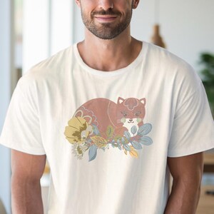 May include: White crew neck t-shirt with a cat and floral design. The cat is brown with a white face, and the flowers are yellow, blue, and green. The design is a stylized illustration. The shirt is a standard fit.