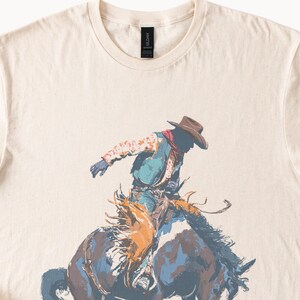 May include: Cream-colored t-shirt featuring a cowboy riding a bucking bronco. The cowboy is wearing a brown hat, a red neckerchief, a patterned shirt, and a blue vest. The horse is depicted in shades of blue, brown, and orange.