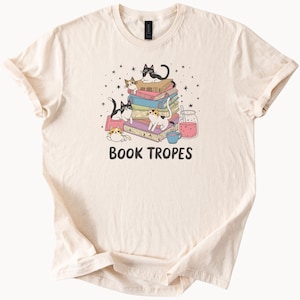 May include: Cream-colored t-shirt featuring a whimsical illustration of several cats lounging on a stack of colorful books. The design includes a drink, stars, and the text "BOOK TROPES" in a playful font. A charming design for book lovers.